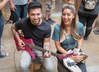 (L–R) Randy Perea and Mireya Medina take a look at Rob Gray’s pointy guitars. After playing, what does Perea like? “The color, of course.” Gray builds multi-scale and extended-range seven- and eight-stringed guitars, and he likes ’em colorful and pointy.