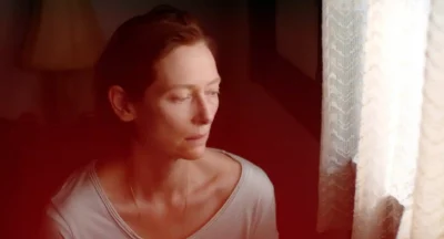 A woman (Tilda Swinton) looks out a window with translucent curtains. She looks sullen. 