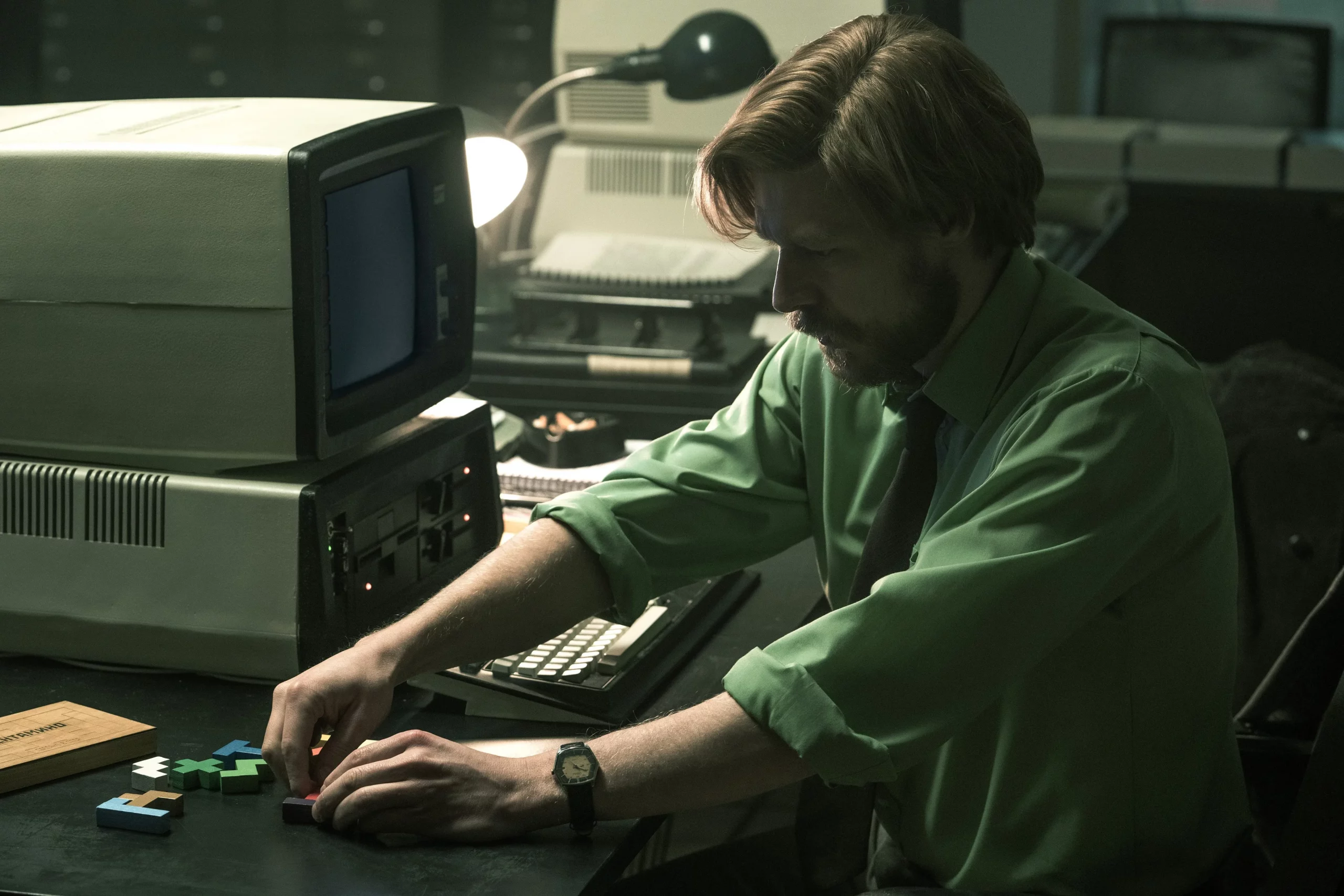 A man sits in a dimly lit office next to an old computer. It's the '80s. He is moving tetriminoes around on a desk.
