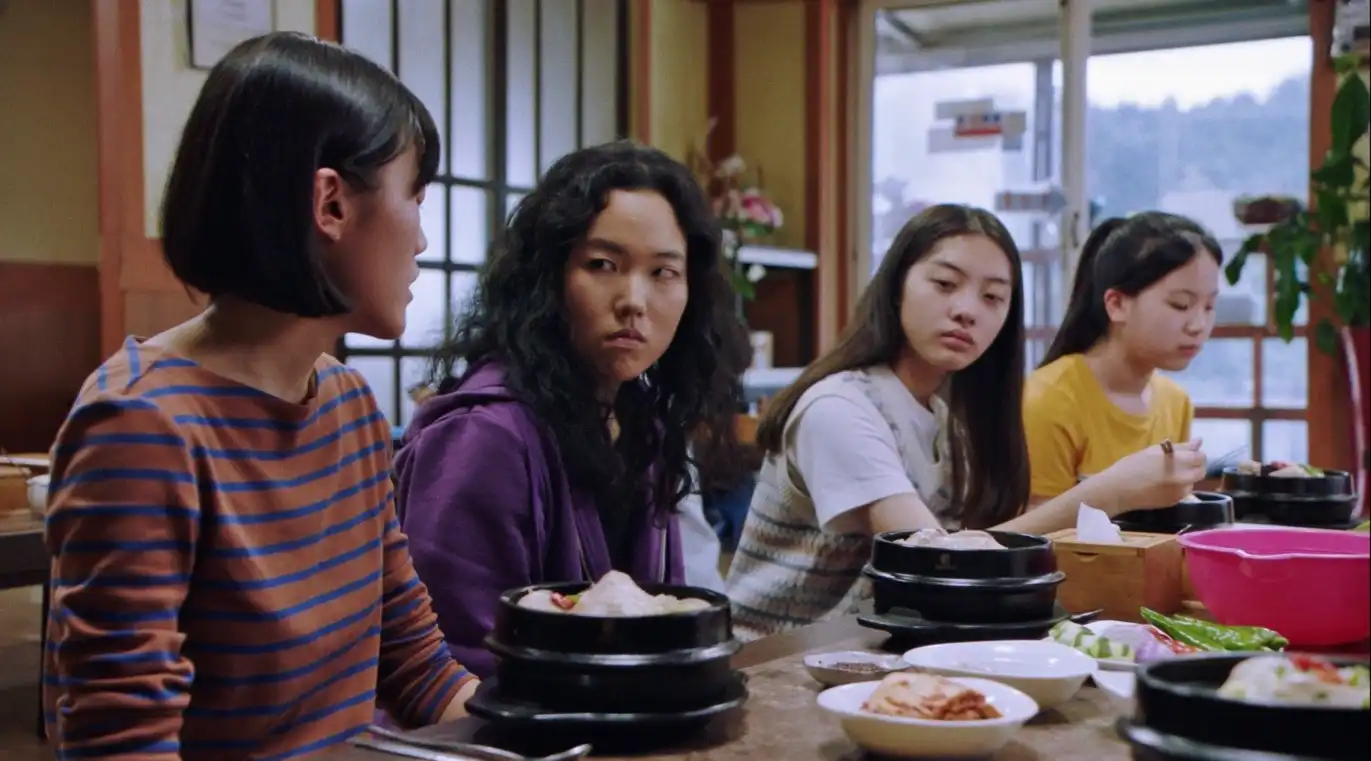Four Korean people sit at a table eating their own meals. They are at a restaurant together. The leftmost person looks at the person to their left, who is looking back at them sullenly.