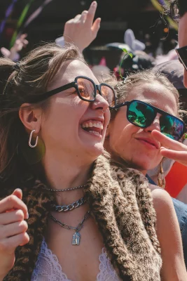 Picture shows two people smiling. Both are wearing large sunglasses. The one in the front has on a fuzzy cheetah-print vest.