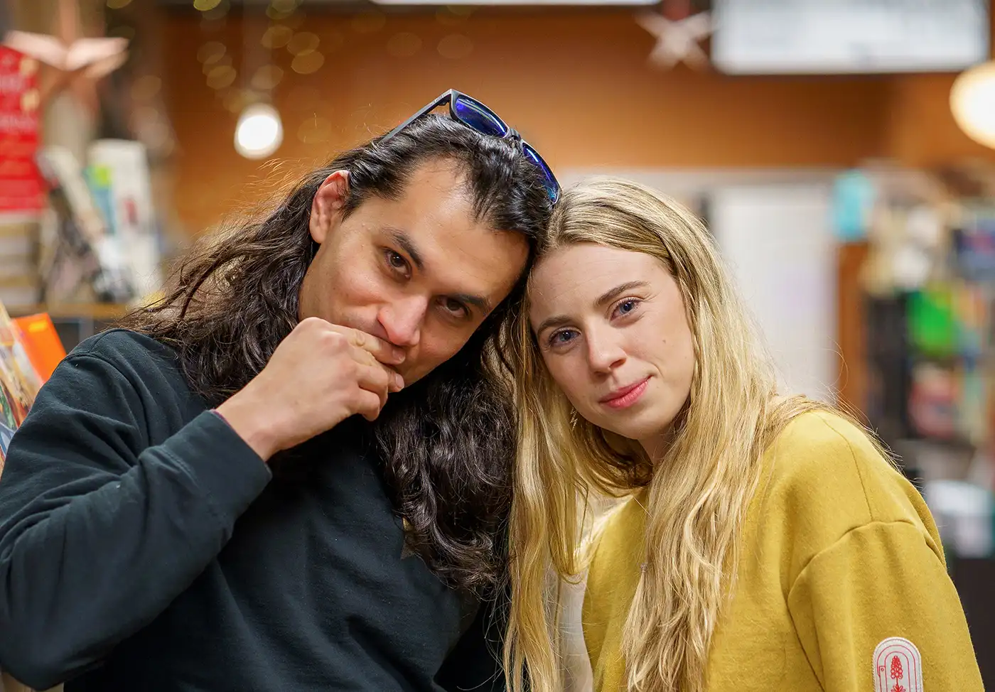 (L–R) Danny Patiño and Bri McCall make up the indie duo The Alpines. In this photo they are standing in a library. Danny leans into Bri, his hand covering his mouth coyly.