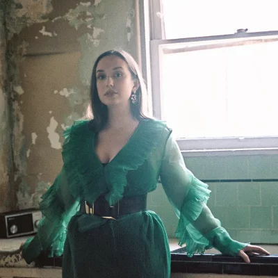 Brooke Trantor stands next to a window with light shining in. She is wearing a a green and billowy outfit with a large black belt. 