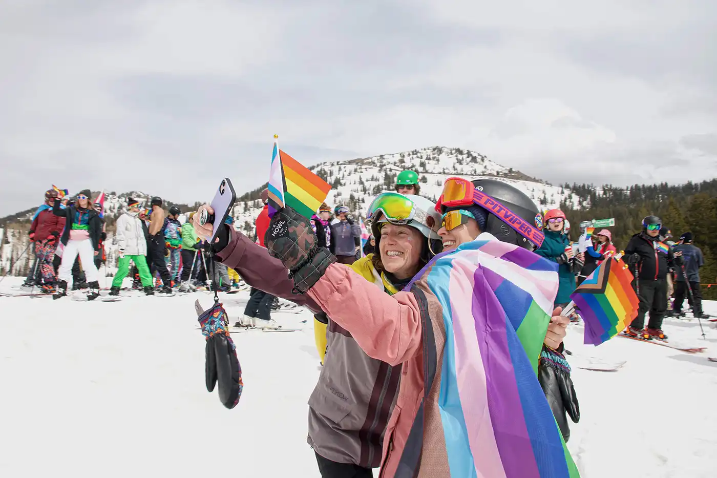 Two women (C Meyer and Michelle Anklan) stand bundled up in winter weather taking a selfie together. They are draped in pride flags. Behind them is a crowd of snow sports enthusiasts.