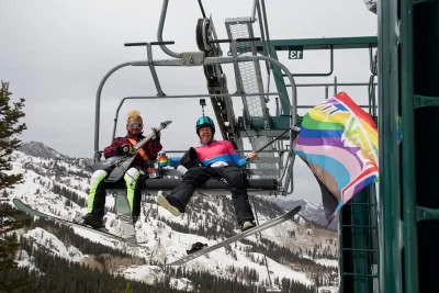 Two skiers ride a ski lift at Brighton Ski Resort on Pride Ride Day 2022. One person is holding a stylish pride flag. Photo: Jovvany Villalobos 