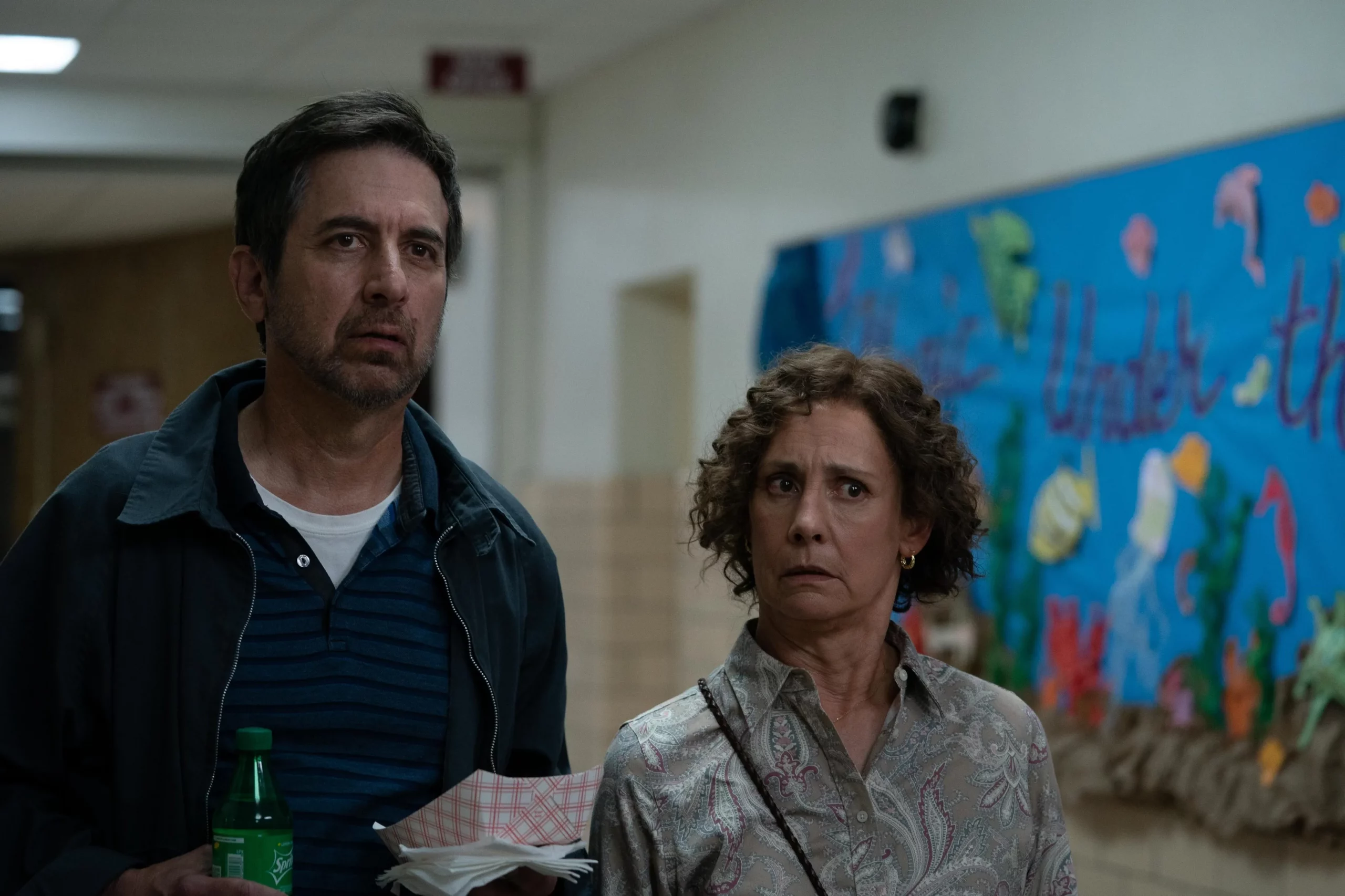 Two adult people give a confused look toward someone off camera. They are in a high school hallway.