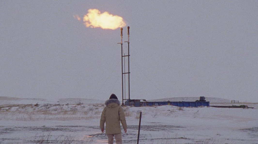 A person is standing in a field looking at a smoke stack–esque pipe structure out of which fire is erupting. It's winter, and the person is bundled up.