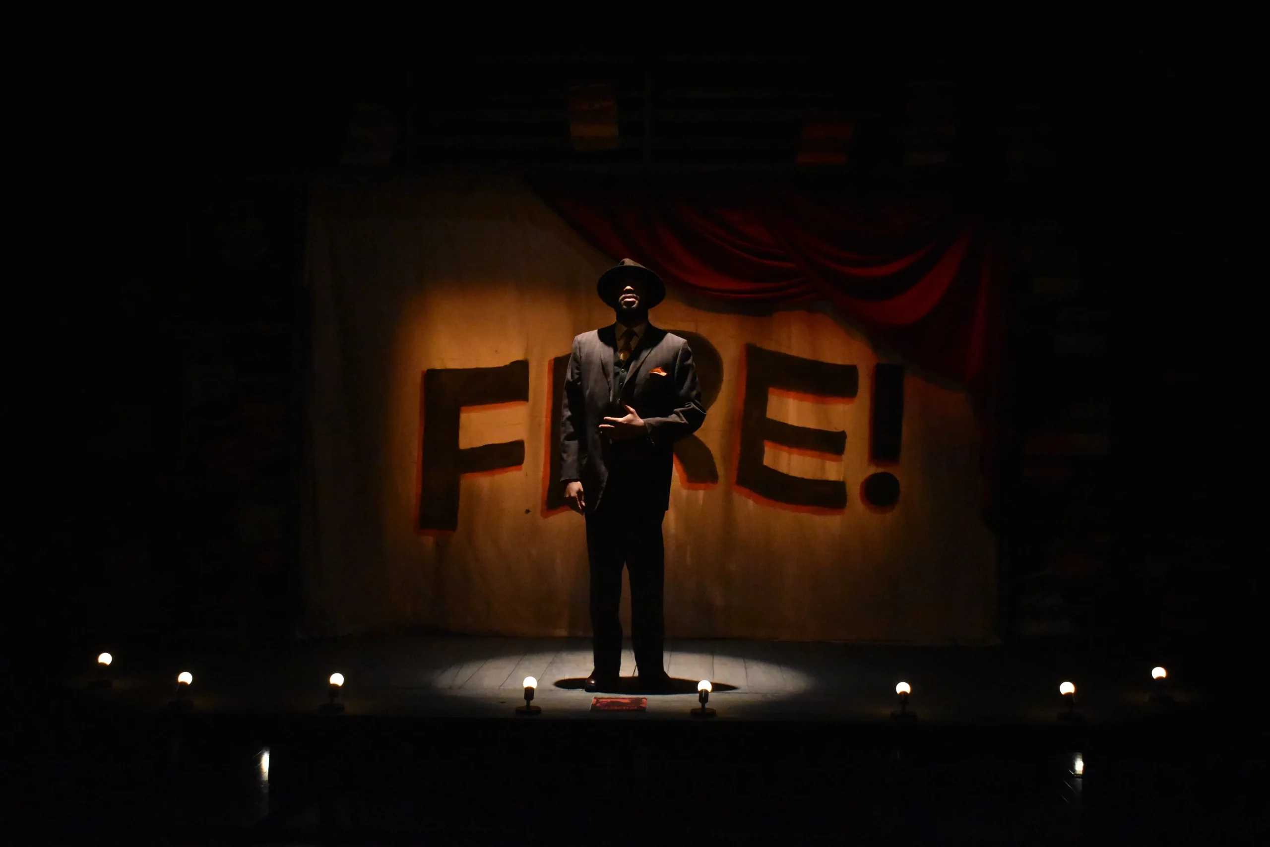 A man stands on center stage shrouded in dark. Behind him read the words, "Fire!"