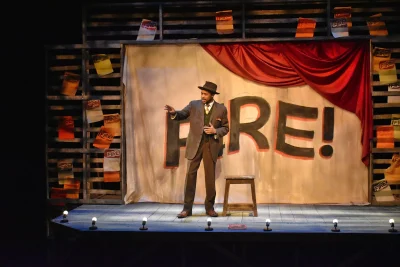 A pulled back shot of a man standing on stage, gesturing toward the audience. Behind him is a curtain that reads, "Fire!"