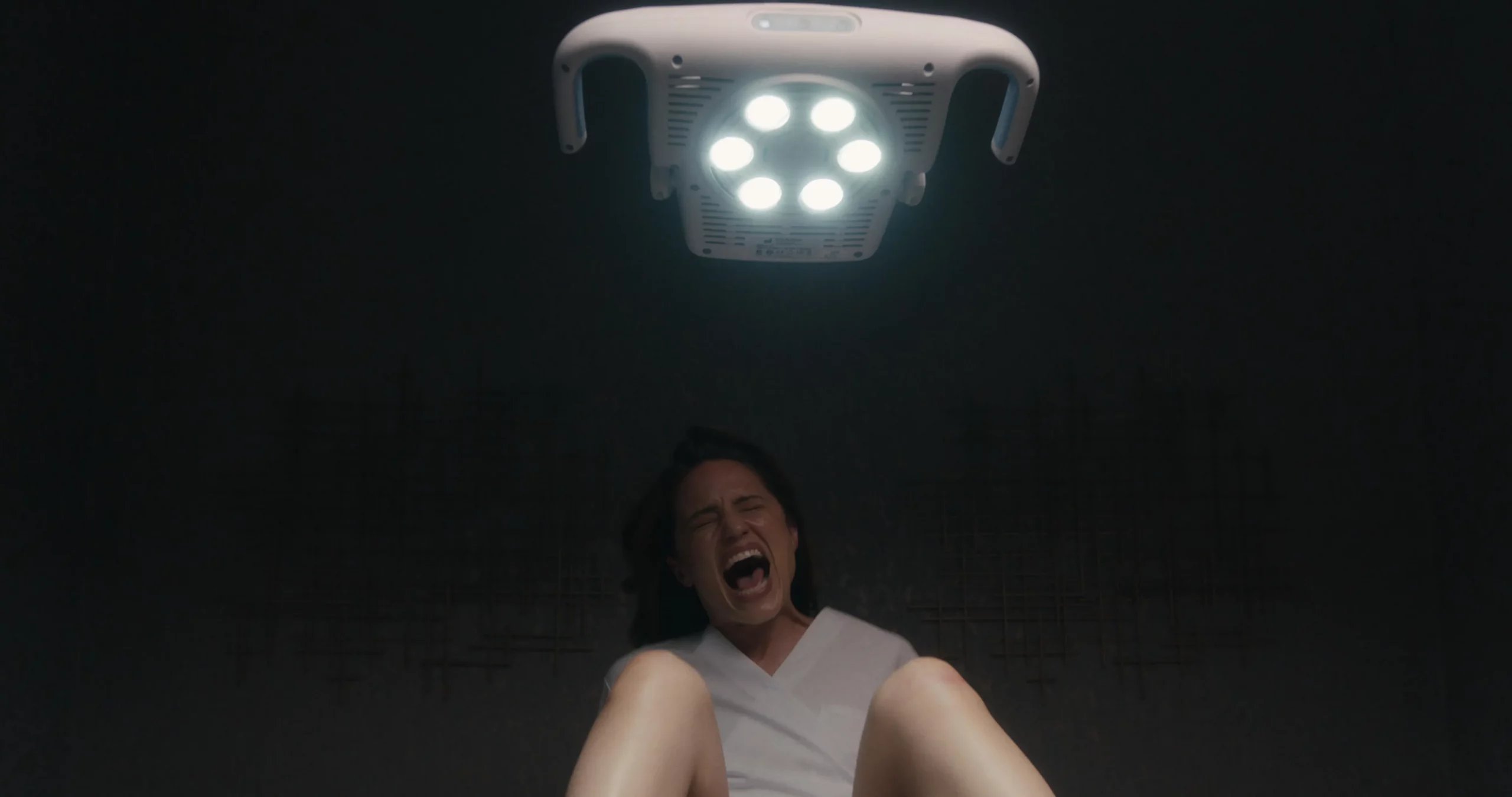 A woman screams in pain underneath a sterile exam room lighting.