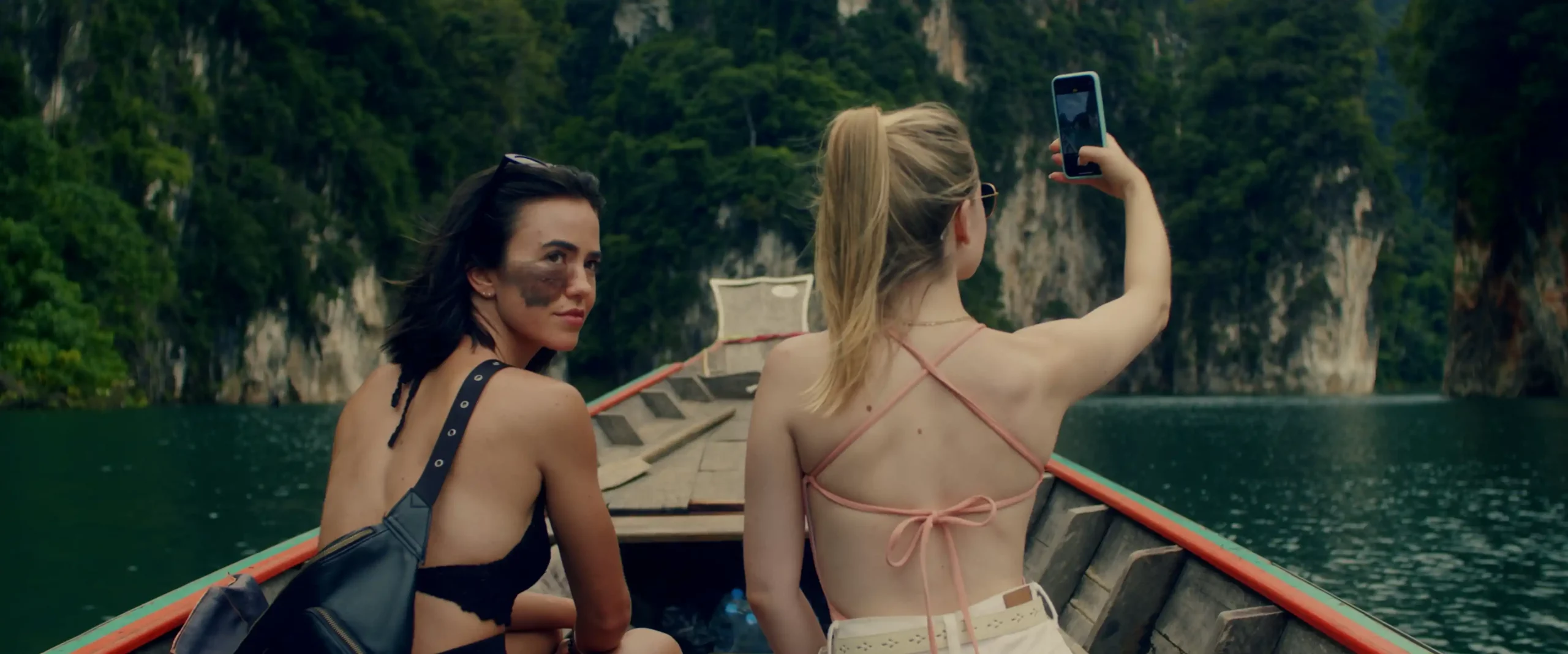 Two girls are on a boat on a river. One of them looks back behind them with a slight smile while the other takes a selfie.