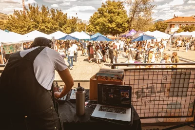 A DJ playing music to the left of the image. behind him a group of tents and Brewstillery-goers.