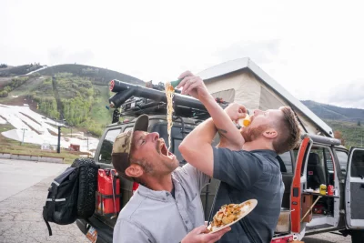 Two men shovel food into their mouths in front of a truck and a mountain range.