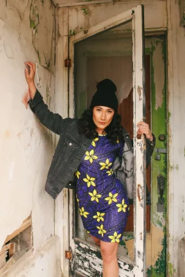 A model gives a serious gaze and wears a dress with flowers, a jacket and a beanie. They stand in front of a dilapidated structure.
