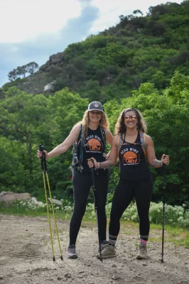 Angelique Fish and Cindy Vance stand together on the trail, carrying backpacks and hiking poles. 