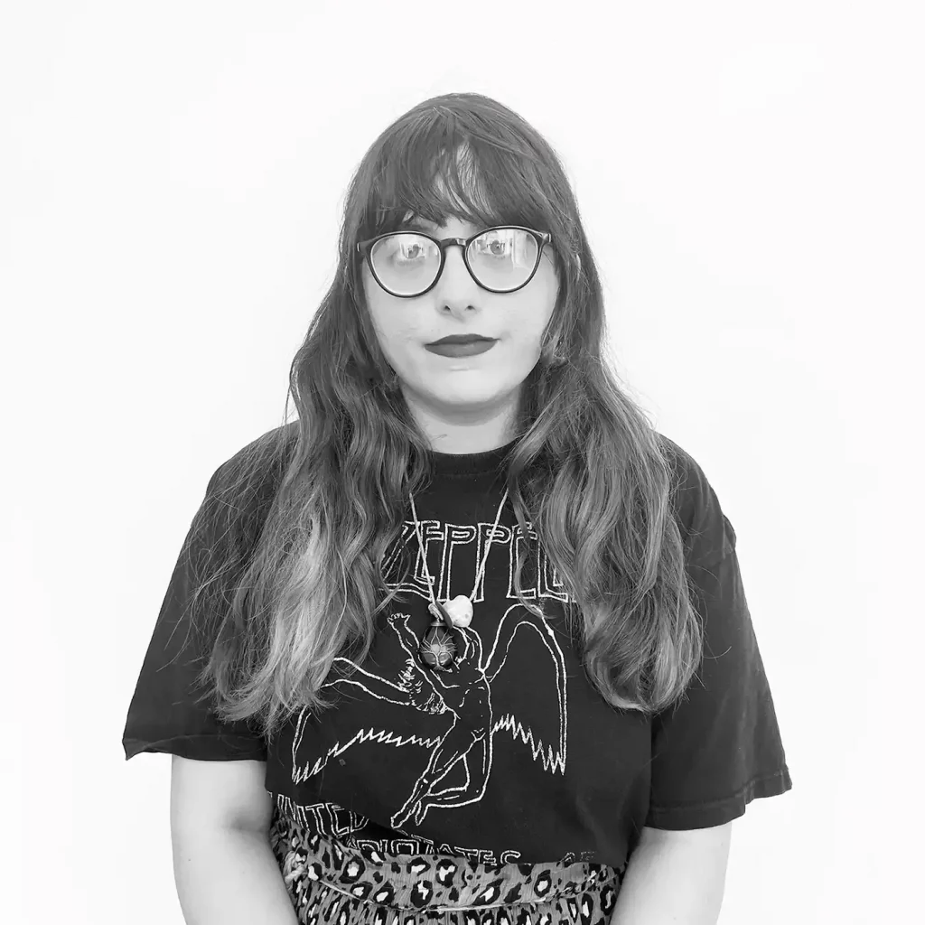 Cherri Cheetah wears a glasses and a black Led Zeppelin shirt, standing in front of a white background.