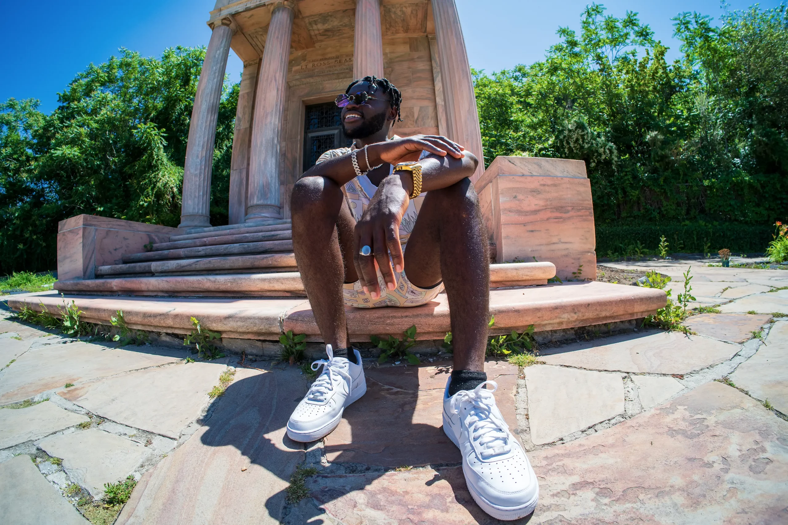 Gavanni sits on the ground in front of a stone structure, wearing white sneakers and gold jewelry.