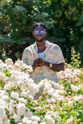 Gavanni looks straight ahead in a field of rose bushes, wearing a light peach shirt and purple-lensed sunglasses. 