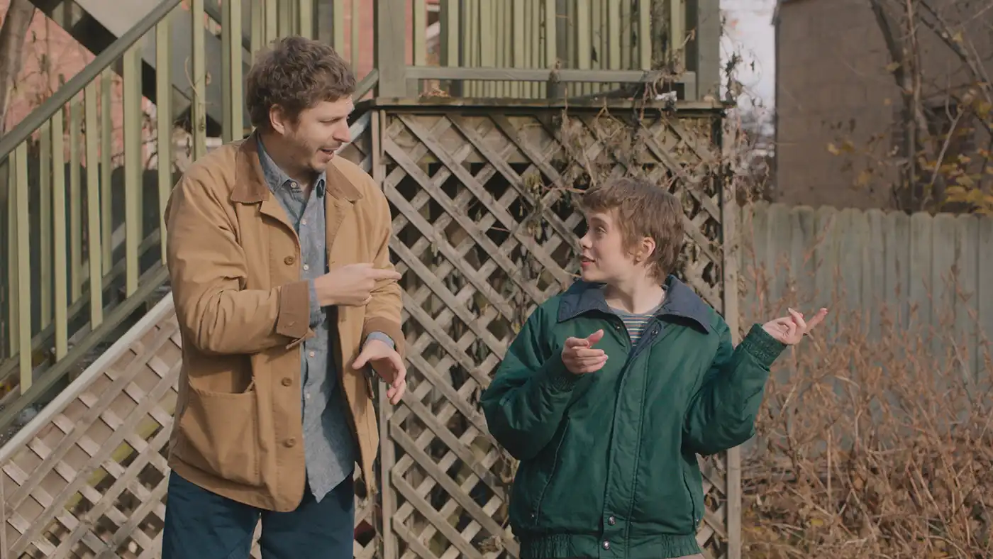 Actors Michael Cera and Sophia Lillis stand in front of a porch staircase, gesturing to one another in conversation.