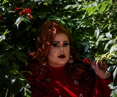 Lexis Monroig stands in red flowy garb adorned with roses against a leafy nature background. Photo: Bonneville Jones