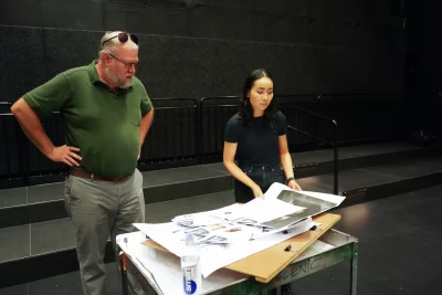 Janice Chan stands in a theater hall looking at planning documents alongside a gentleman in green.