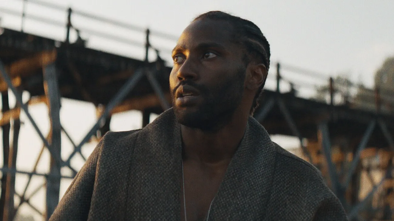 John David Washington in The Creator (2023) stands against a backdrop of a bridge and stares off screen