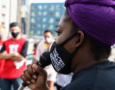 Michelda George speaks to a crowd through a microphone at the Say Their Names Memorial. She is wearing a Black Lives Matter mask.