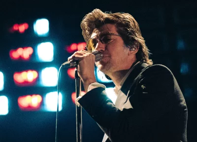 Lead singer for Arctic Monkeys Alex Turner signing into a microphone, wearing yellow sunglasses.