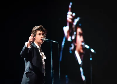 Alex Turner singing into a microphone, with a projects image of himself behind him.