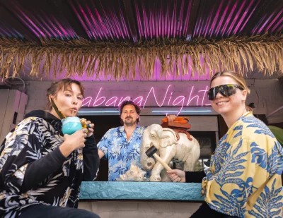 Three people in Hawaiian shirts sit around a table with an elephant on top. One sips a drink. A pink neon sign reads "Cabana Nights" in the background.