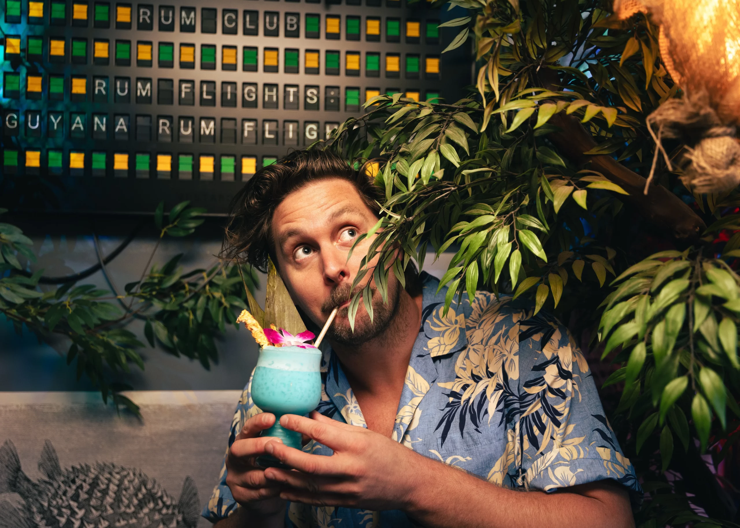 Mikey Edwards wears a Hawaiian shirt and sips from a bright blue cocktail in ACME Bar Co.
