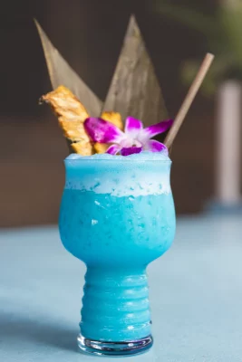 A bright blue rum cocktail garnished with a purple flower and piece of pineapple.