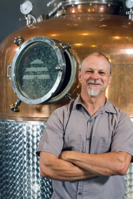 Chris Barlow, one of the co-owners of Beehive Distilling, stands in front of distilling apparatus with a beehive logo.