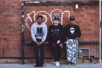 Mal, Saco and Andrew pose in front of a brick wall with graffiti.