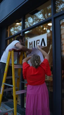 Two people standing outside of the gallery, sticking a window decal reading "MICA" on the window.