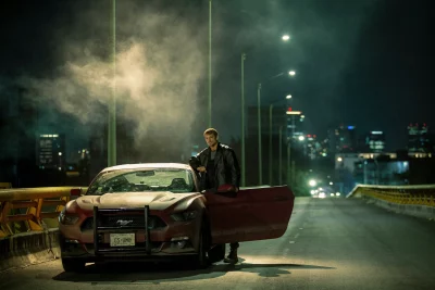 In a still from Silent Night, Joel Kinnaman stands next to a car on a bridge at night.