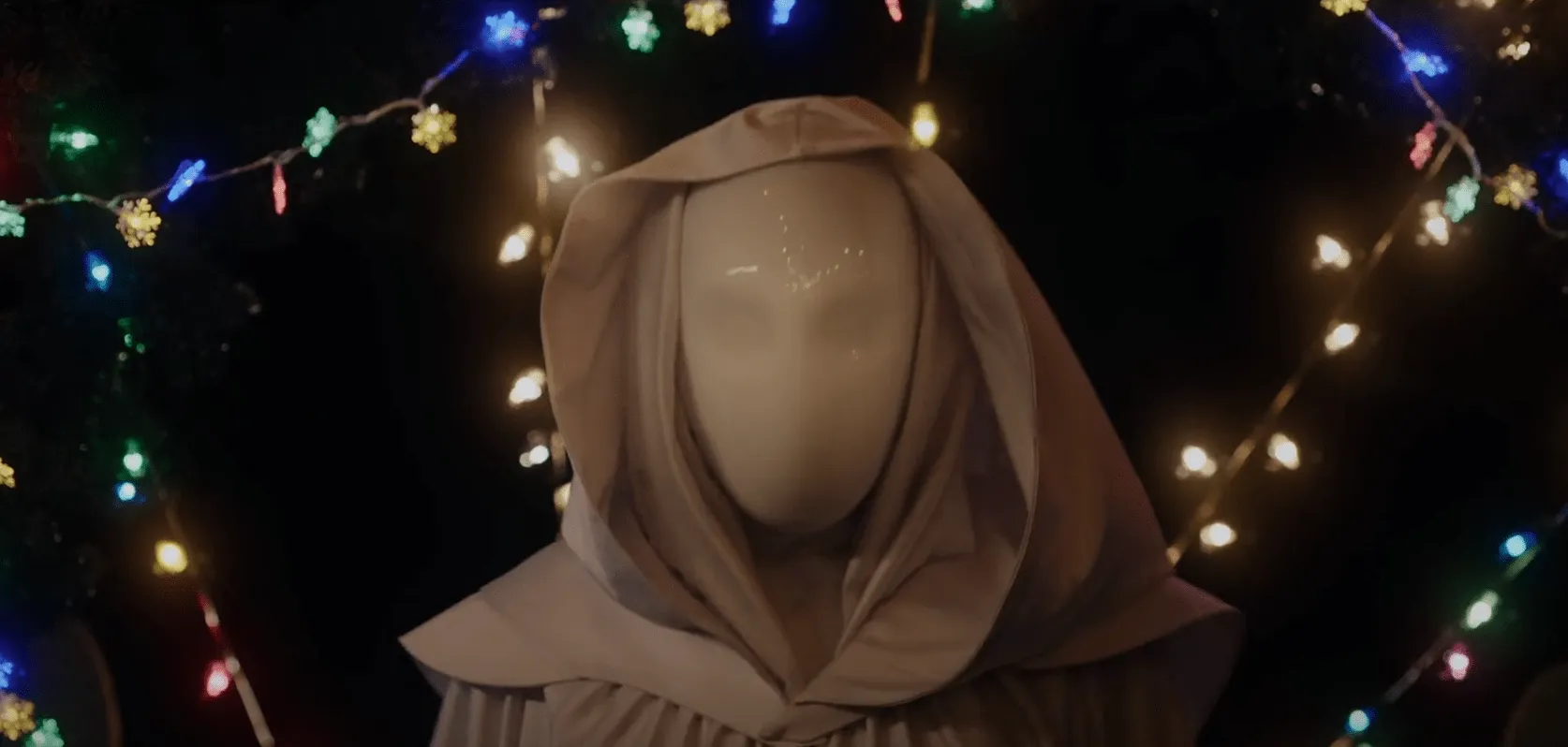 Still from It's A Wonderful Knife. A person with a white mask looks into the camera. Behind them are Christmas lights.