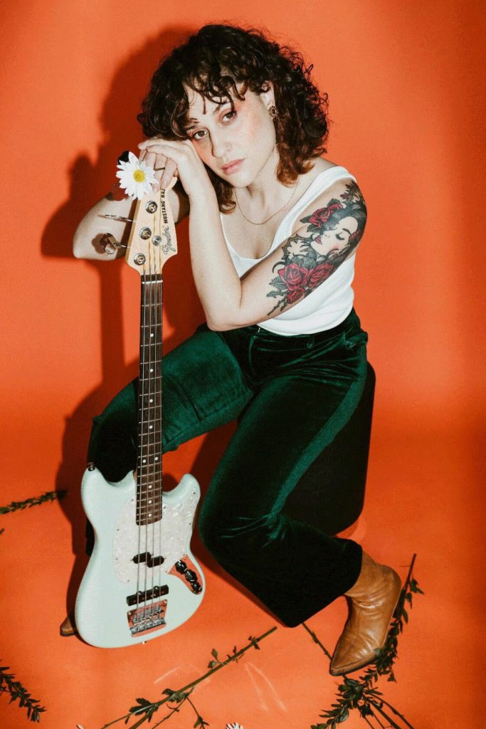 Elowyn poses with her bass guitar in front of an orange background