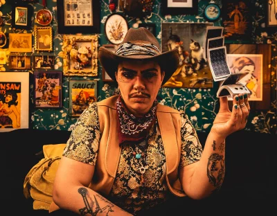 Heller Highwater is dressed like a cowboy in front of a wall covered in pictures, with a deck of cards in hand.