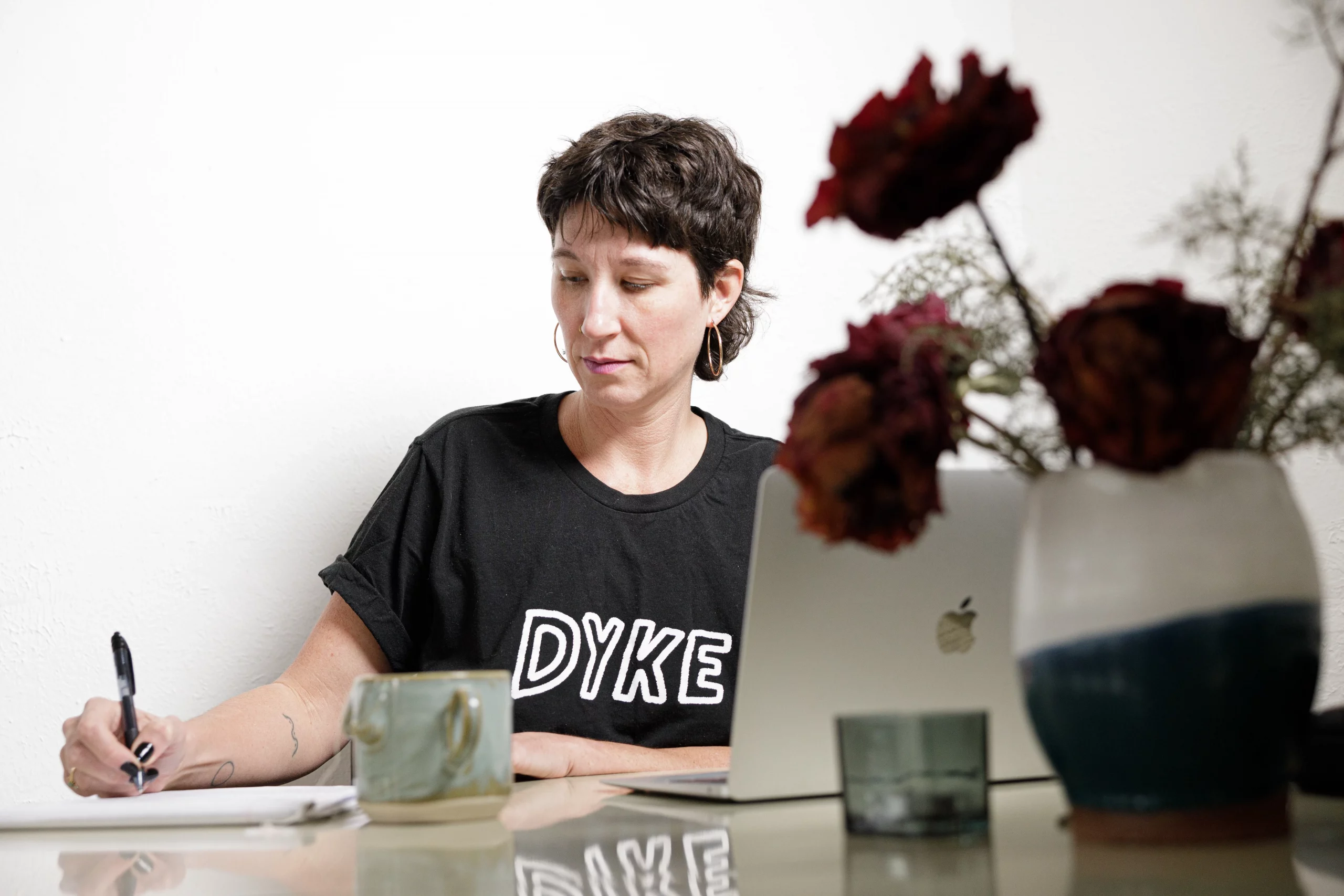 Amanda Madden writes at a table with flowers in front of them, wearing a shirt that reads DYKE.