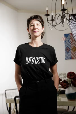 Amanda Madden stands in a dining room, wearing a black shirt that says "Dyke" with their hands in their pockets.