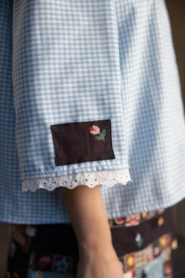One of Jenson's designs where a small black patch with a flower is sewn onto the sleeve of a blue gingham blouse.