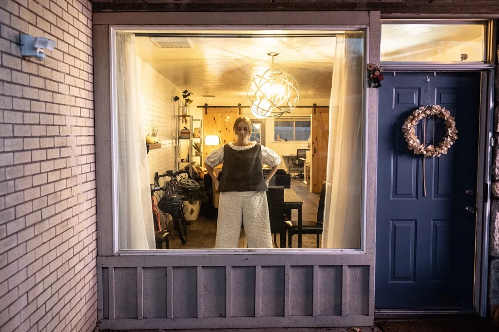 Abigail Rue Jenson stands in her home, visible through a window next to the front door. She is standing with her hands at her hips and is wearing bloomers and a boxy brown and white blouse.