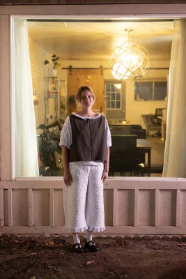 Abigail Rue Jenson stands in front of a window to her home in a boxy blouse and bloomers.