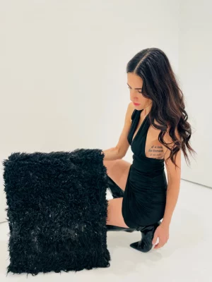 Samantha DaSilva, in a black mini dress, sits next to her framed black sheepskin piece.
