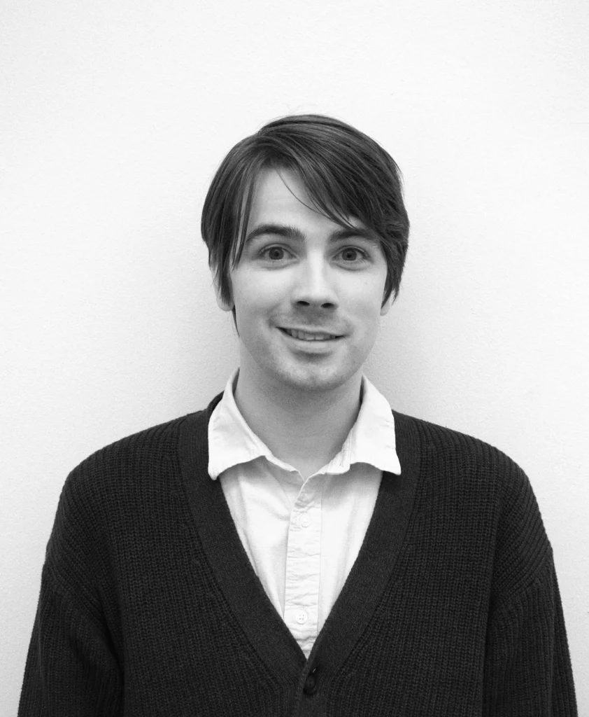 A black and white photo of Daniel Kirkham, wearing a button down and a cardigan.