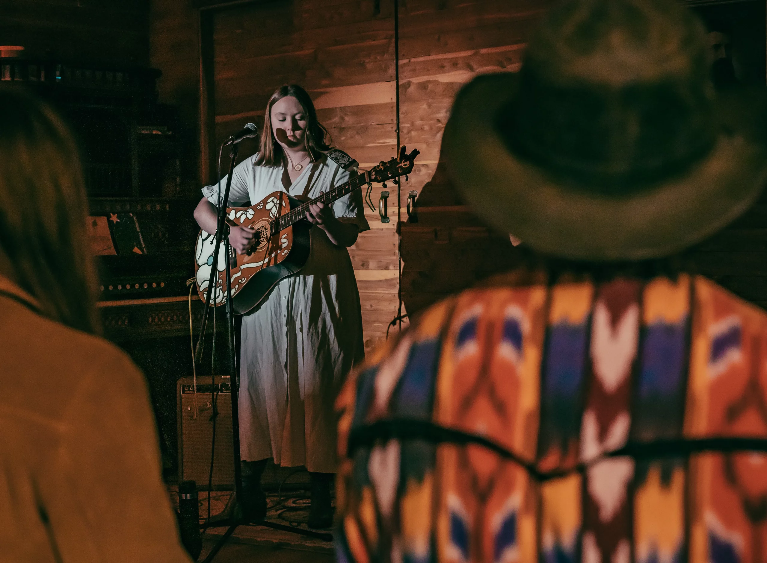 A young woman in a white dress stands in a wood paneled room, holding a guitar and singing into a microphone for a crowd.