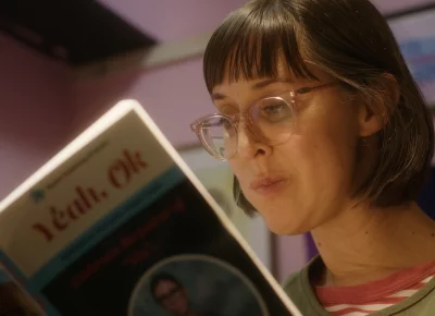A bespectacled woman with short brown hair reads a book in a pink-walled bedroom.