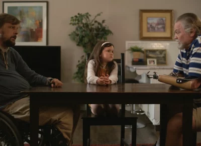 Two middle aged men sit on opposite ends of a kitchen table with a young girl between them.