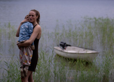 A woman holds her child in front of a lake.